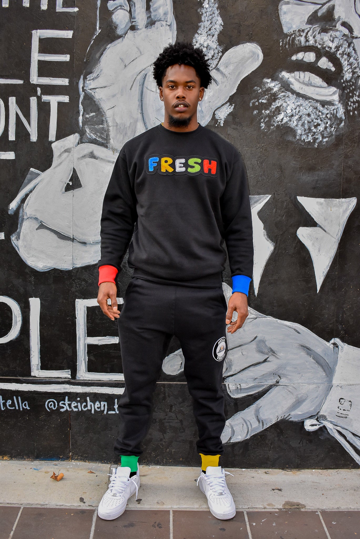 Model wear black crewneck sweatshirt with fresh center chest logo. Sweatshirt has one red cuff and one blue cuff. Sweatpants have one green cuff and one yellow cuff.