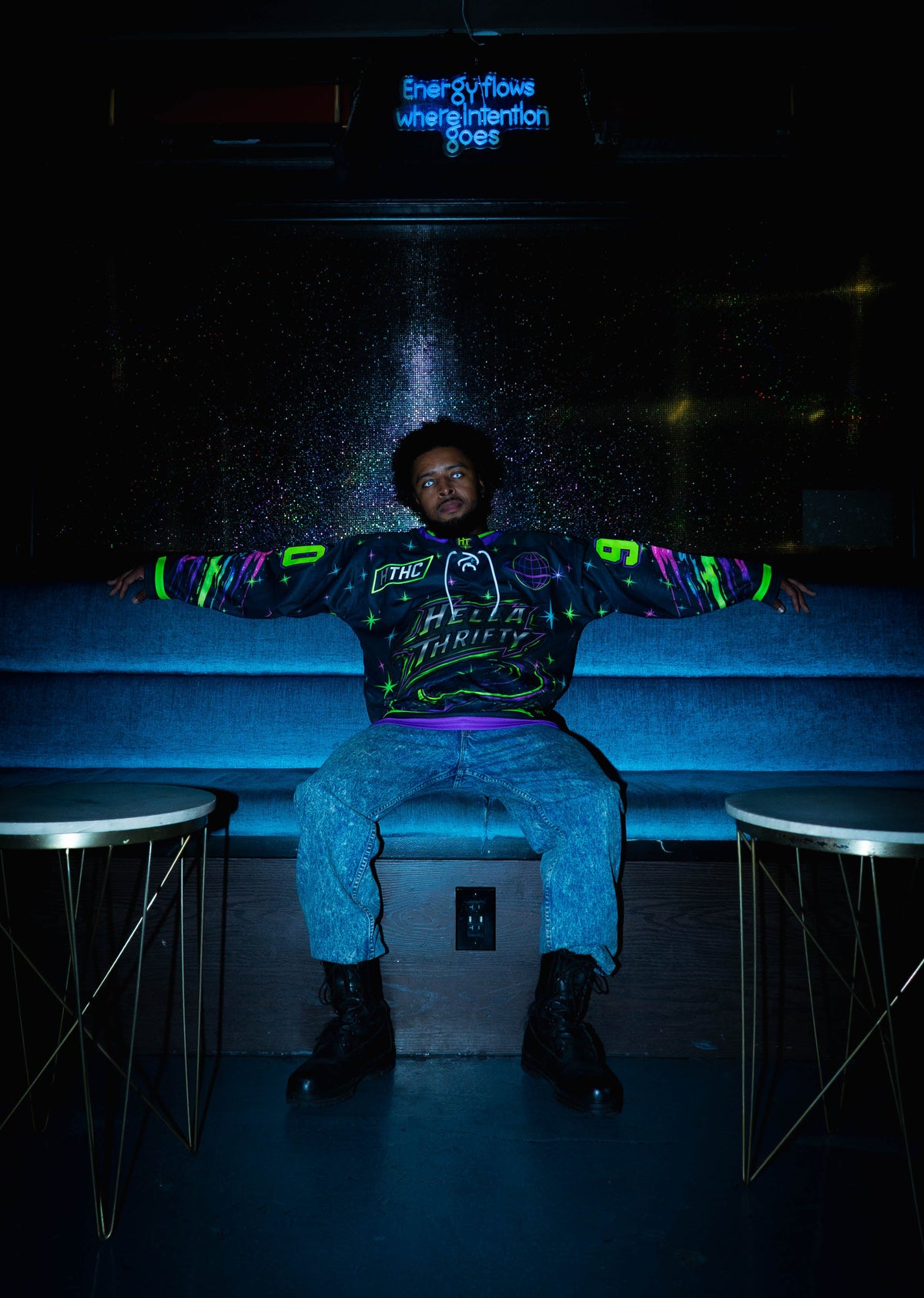 A man wearing a black graphic UV hockey jersey with neon green and purple accents, blue jeans, and black boots sits on a blue couch in a dark room with a glowing blue sign in the background.