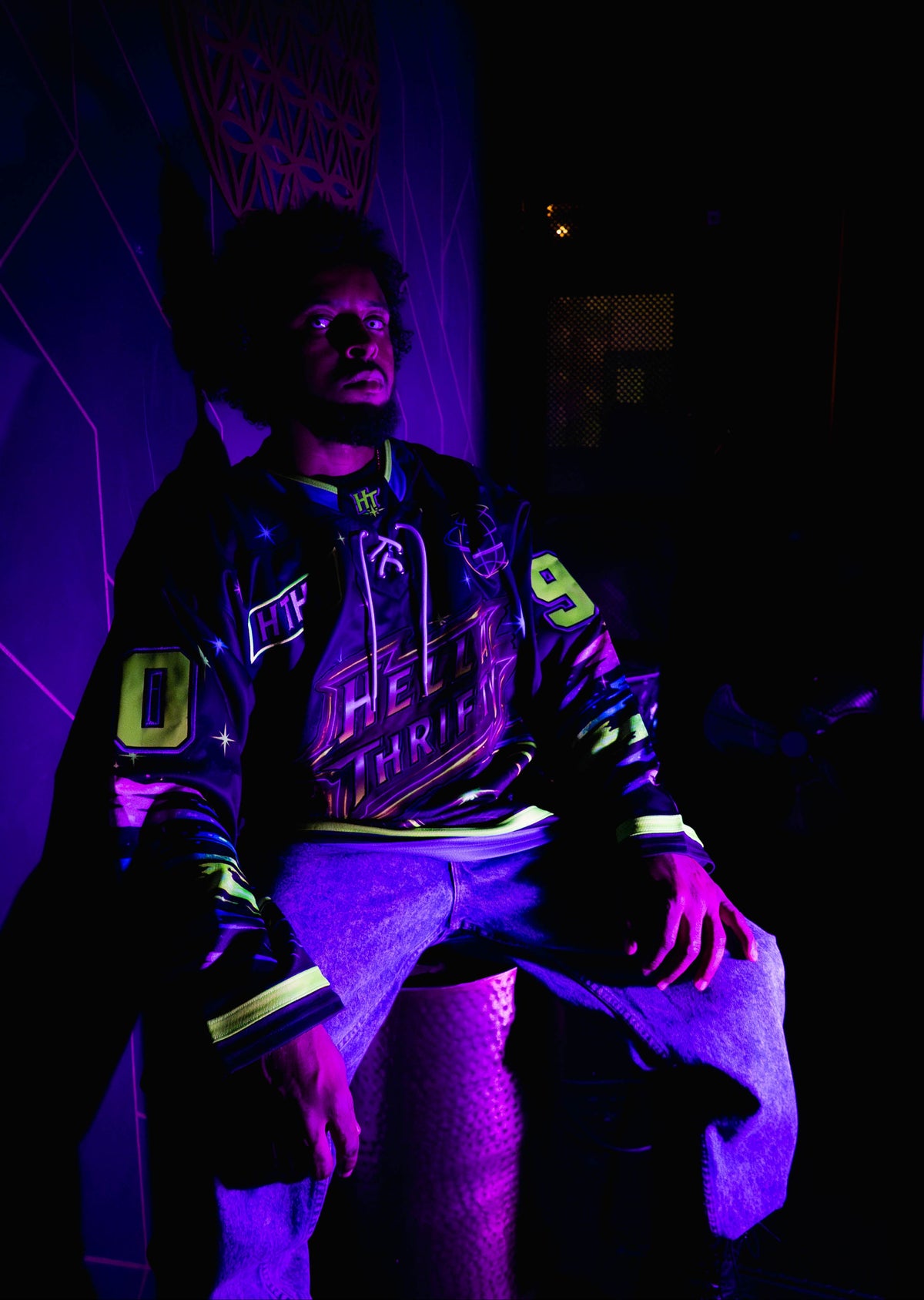 A person wearing a uv hockey jersey with neon green and purple lettering, posed in a dark room under UV-like lighting that casts strong purple and pink shadows.